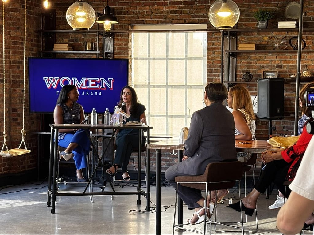 Louise Jones and Kalisha Figures speaking at the Women for Alabama Fireside Chat, seated at a table on a stage in front of a "Women for Alabama" digital screen.
