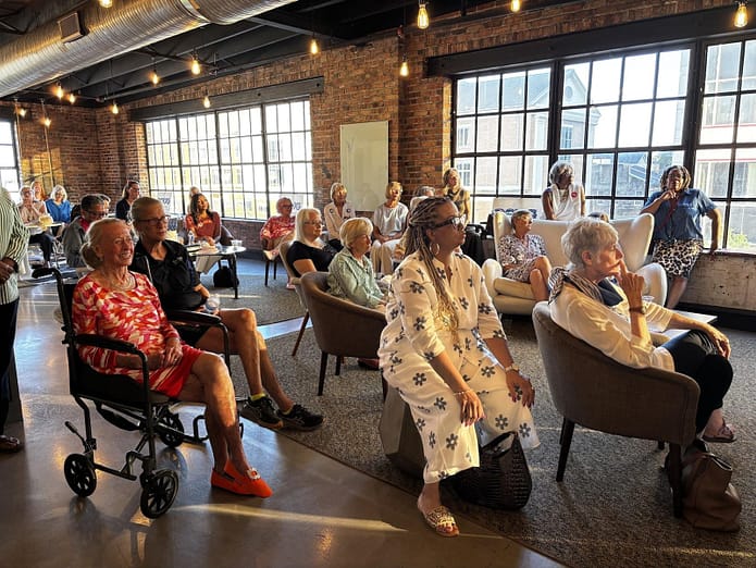 Attendees seated in chairs and a wheelchair, listening intently during the Women for Alabama Fireside Chat event in a room with exposed brick and large grid windows.