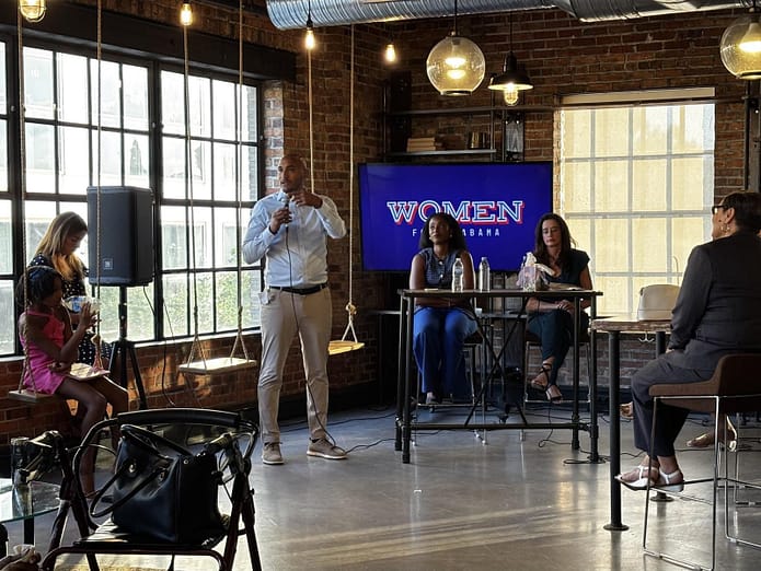 Second Congressional District candidate Shomari Figures speaking into a microphone at the Women for Alabama Fireside Chat event, with Louise Jones and Kalisha Figures seated at a table on stage.